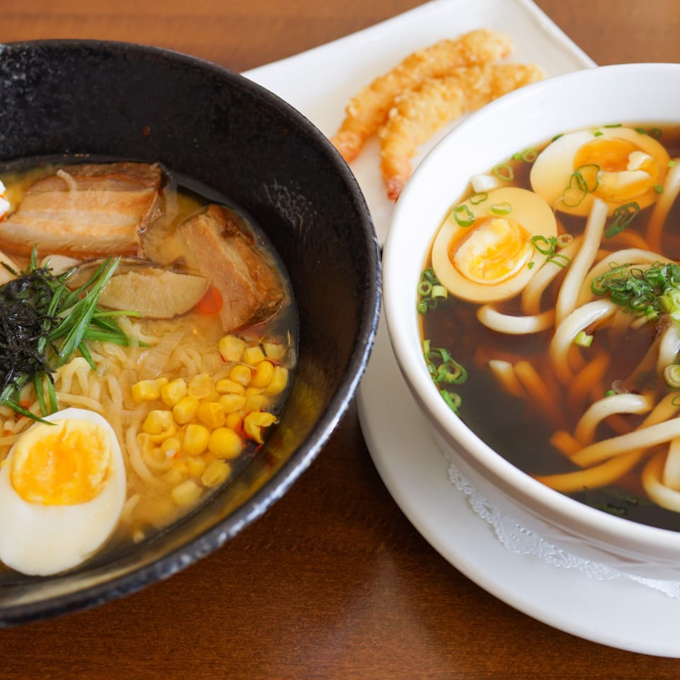 Two bowls of ramen and udon with eggs, corn, and pork, alongside fried shrimp tempura.