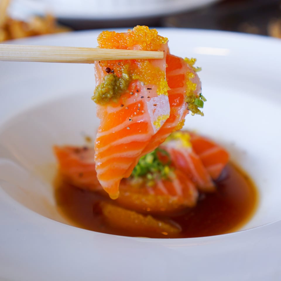Chopsticks holding a piece of salmon sashimi with roe and green garnish over a bowl of sauce.