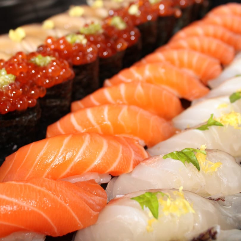 Rows of fresh sushi, including salmon nigiri, salmon roe gunkan, and white fish nigiri with garnishes.