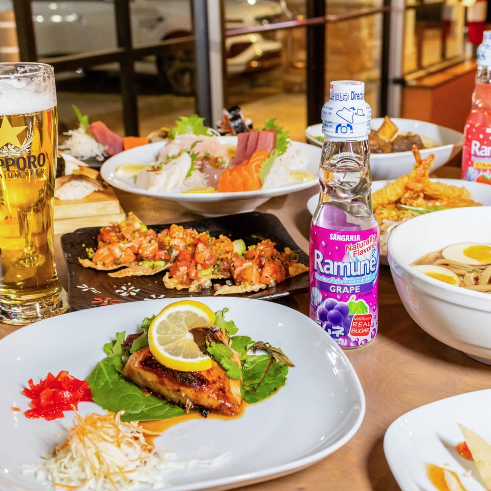 A table laden with various Japanese dishes, including sushi, ramen, grilled fish, and drinks.