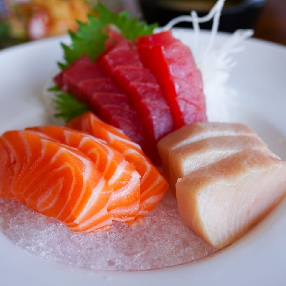 A white plate holds fresh salmon, tuna, and white fish sashimi on a bed of ice.