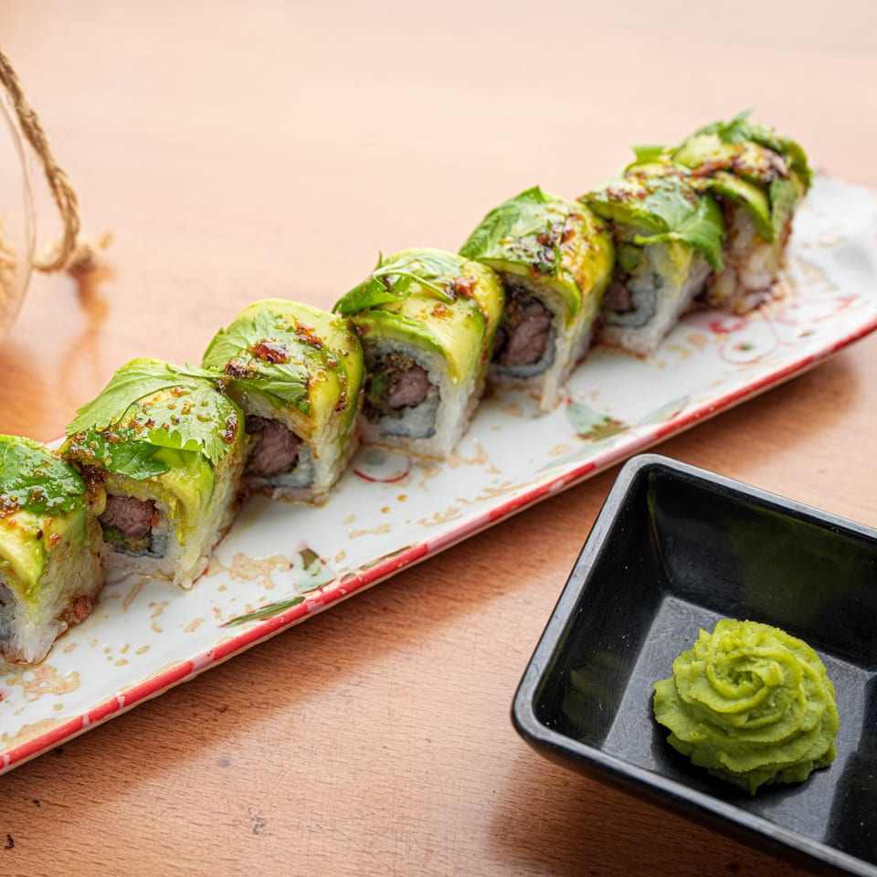 Sushi rolls with avocado, herbs, and sauce on a decorative plate, with wasabi in a black dish.