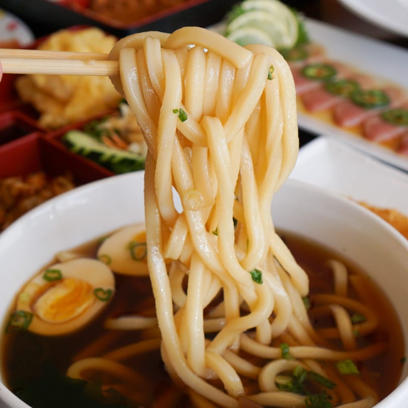 Chopsticks lifting thick udon noodles from a bowl of broth with sliced eggs and scallions.
