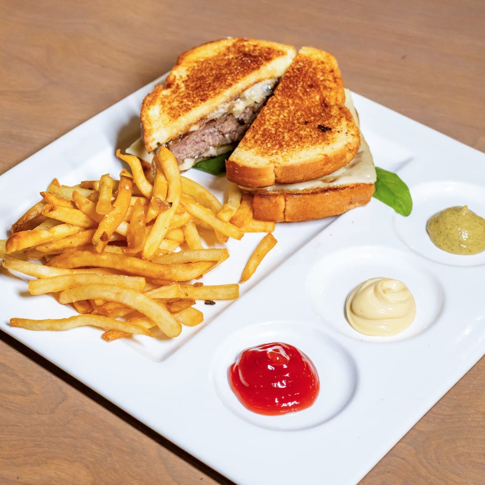 Grilled cheese sandwich with a beef patty, fries, ketchup, mayonnaise, and mustard on a white plate.