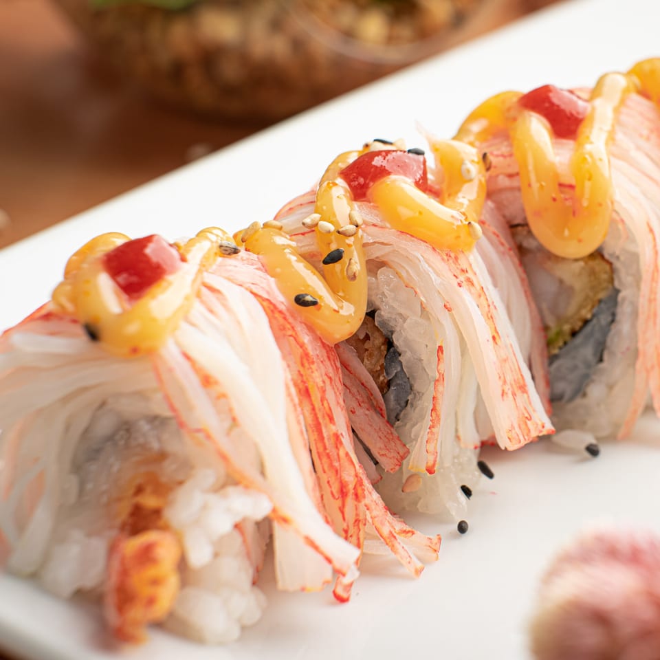 Sushi roll topped with crab meat, yellow sauce, red sauce, and sesame seeds on a white plate.
