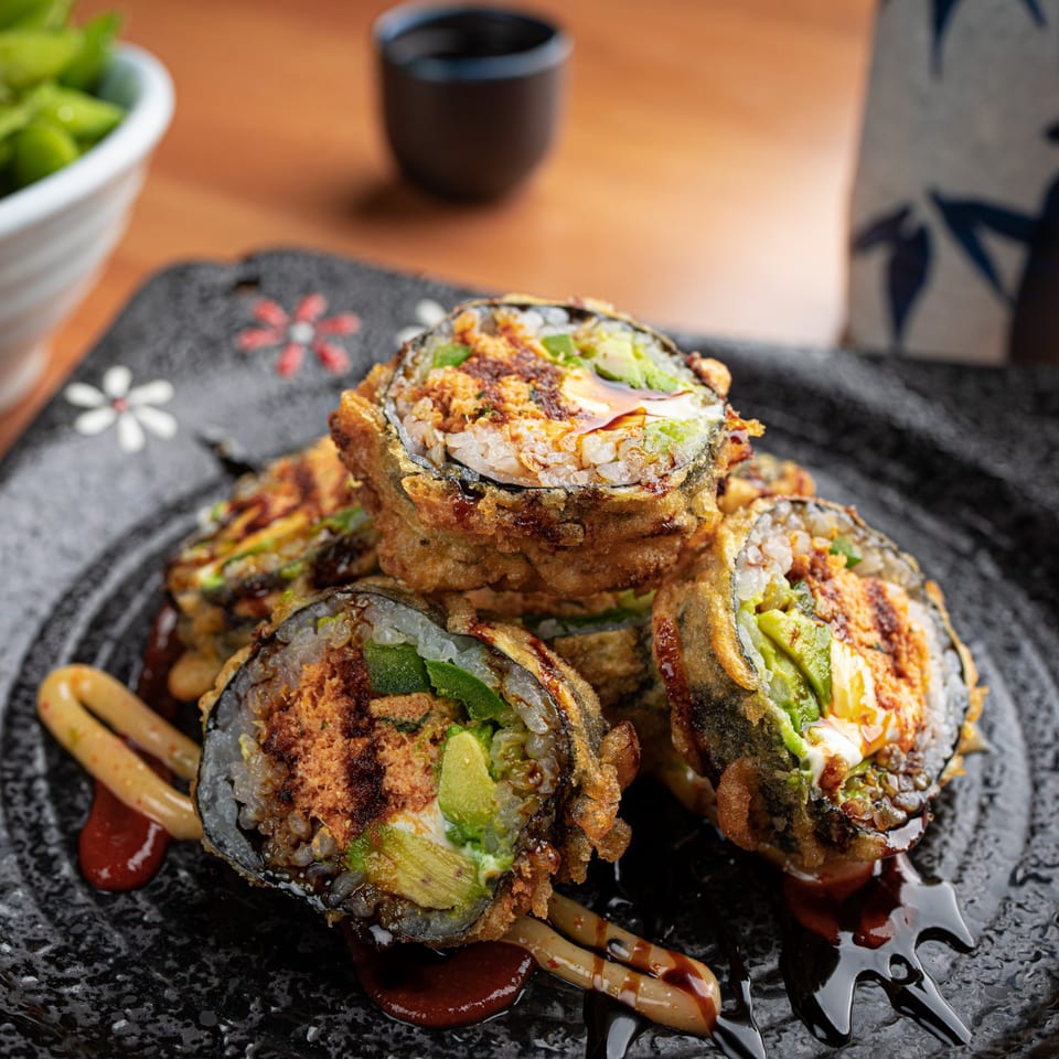 Crispy fried sushi rolls with avocado, sauce, and toppings on a decorative black plate.