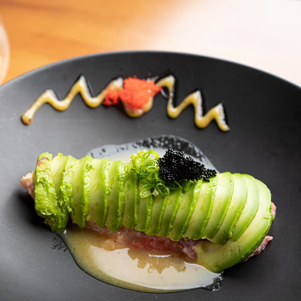 Avocado-wrapped sushi roll topped with black caviar and green onions on a black plate.